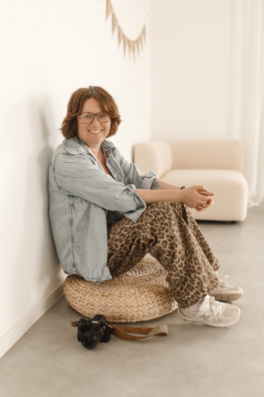 Fotografin in lässigem Denim-Outfit sitzt entspannt auf einem Sitzkissen vor einer weißen Wand.